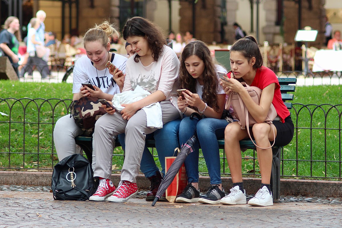 Four young women sit on a park bench, each engrossed in their smartphones, illustrating how technology can change social interaction. The image highlights the theme of Sherry Turkle's work on being "Alone Together."
