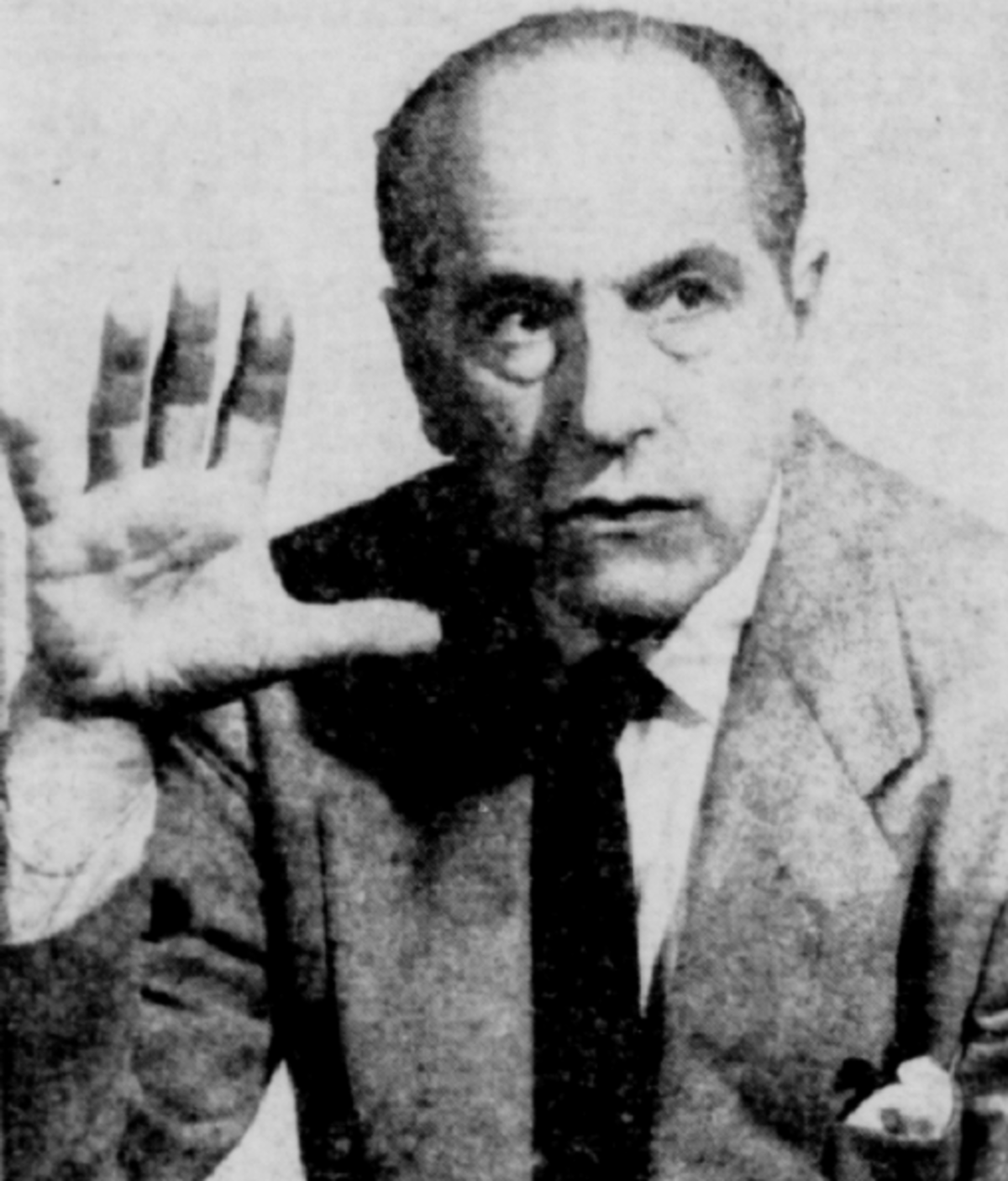 Black and white portrait of mentalist Joseph Dunninger from 1956, looking intently with his right hand raised. He is wearing a suit and tie.