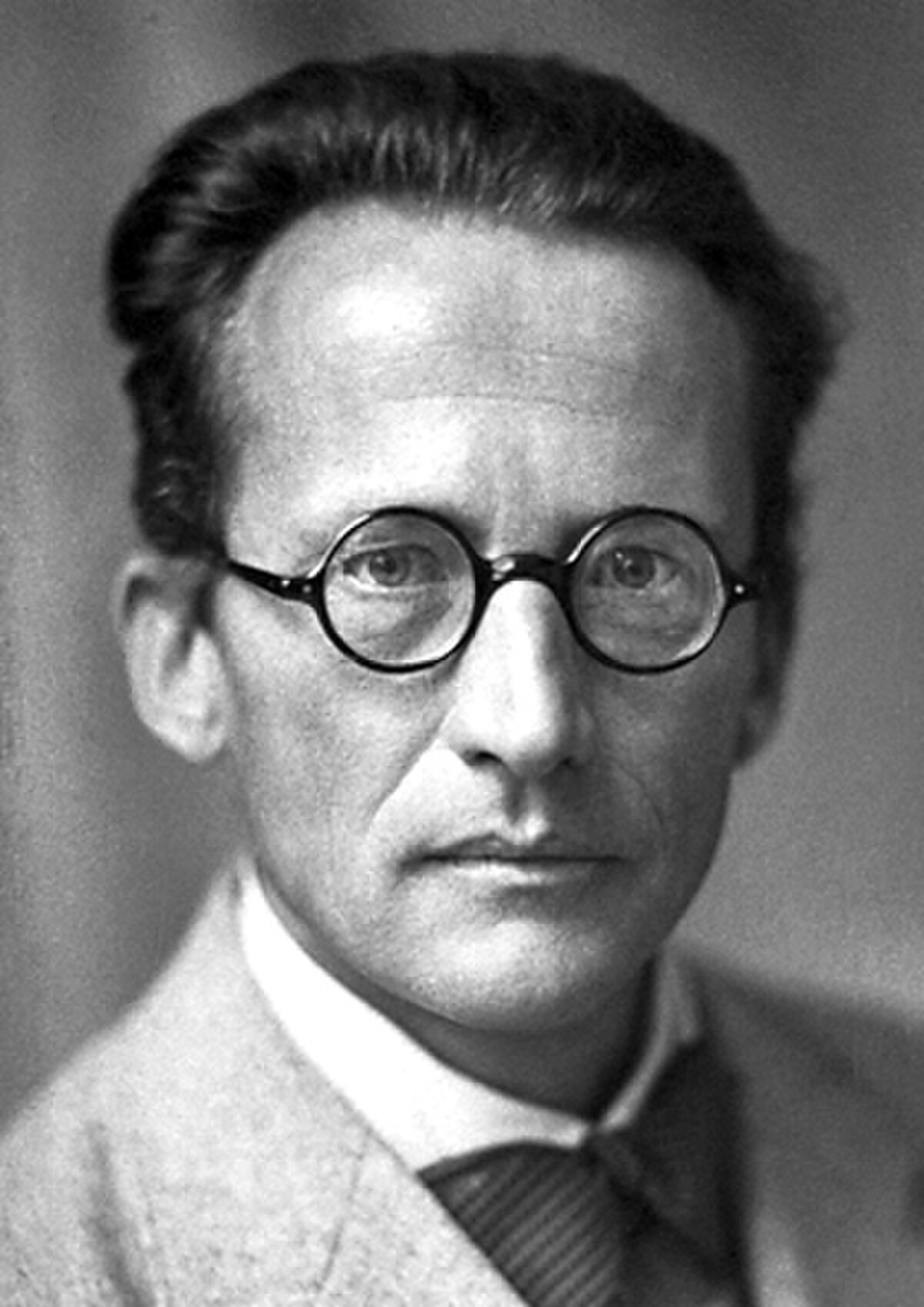 A black and white portrait of Erwin Schrödinger, taken in 1933, shows him wearing round glasses, a suit jacket, and a tie. He looks directly at the viewer with a serious expression.