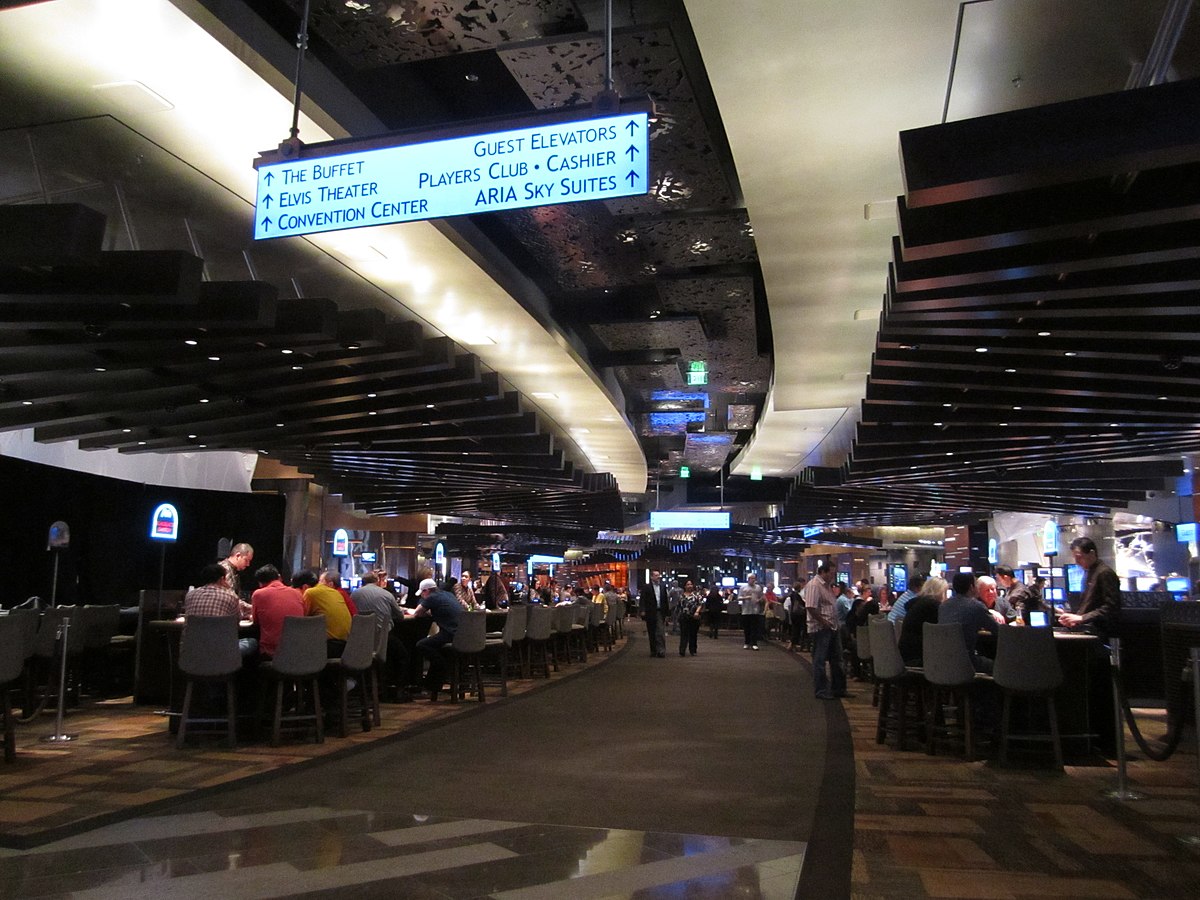 The casino floor of the Aria Resort and Casino in Las Vegas, with rows of gaming tables and slot machines. Overhead signs direct visitors to "THE BUFFET", "Elvis Theater", "Convention Center", "GUEST ELEVATORS", "PLAYERS CLUB", "CASHIER", and "ARIA Sky Suites".