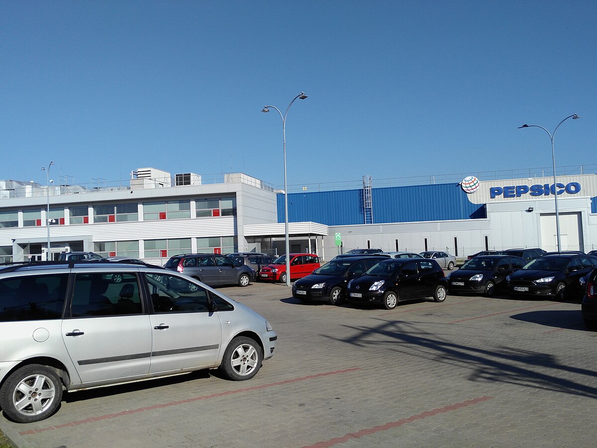 The PepsiCo factory in Tomaszów Mazowiecki, Poland, where Cheetos are produced, featuring a modern building with a blue section and the PepsiCo logo, surrounded by a parking lot with several cars.