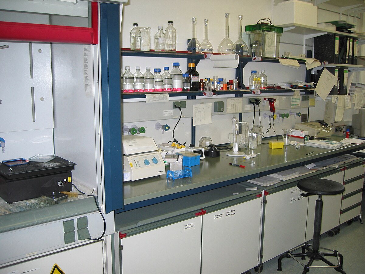 A laboratory bench at the Institute of Biochemistry, University of Cologne in Germany, equipped with various glassware, bottles of reagents, and scientific instruments. The bench features a fume hood on the left and shelves filled with supplies.