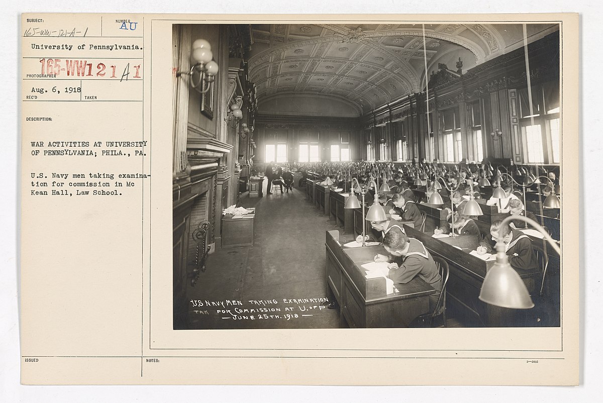 University of Pennsylvania students taking a United States Navy examination for commission in McKean Hall at Penn Law in June 1918. The large hall is filled with rows of students in uniform seated at individual desks with lamps, taking an exam.