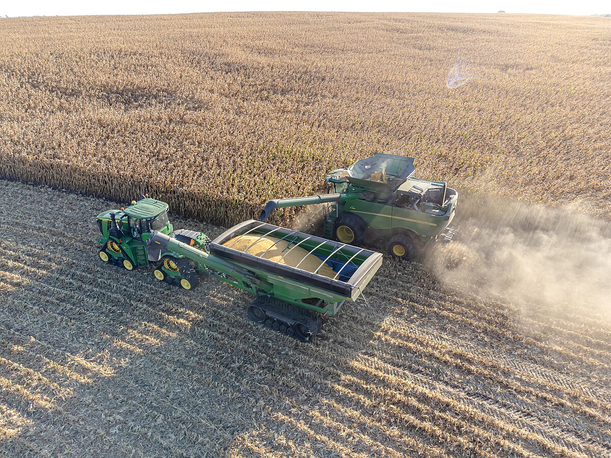 Aerial view of a green John Deere combine harvester harvesting corn in a field, with a green grain cart pulled by a tractor alongside it. The combine is offloading harvested grain into the grain cart while both machines move through the field, leaving a trail of dust and cut stalks.