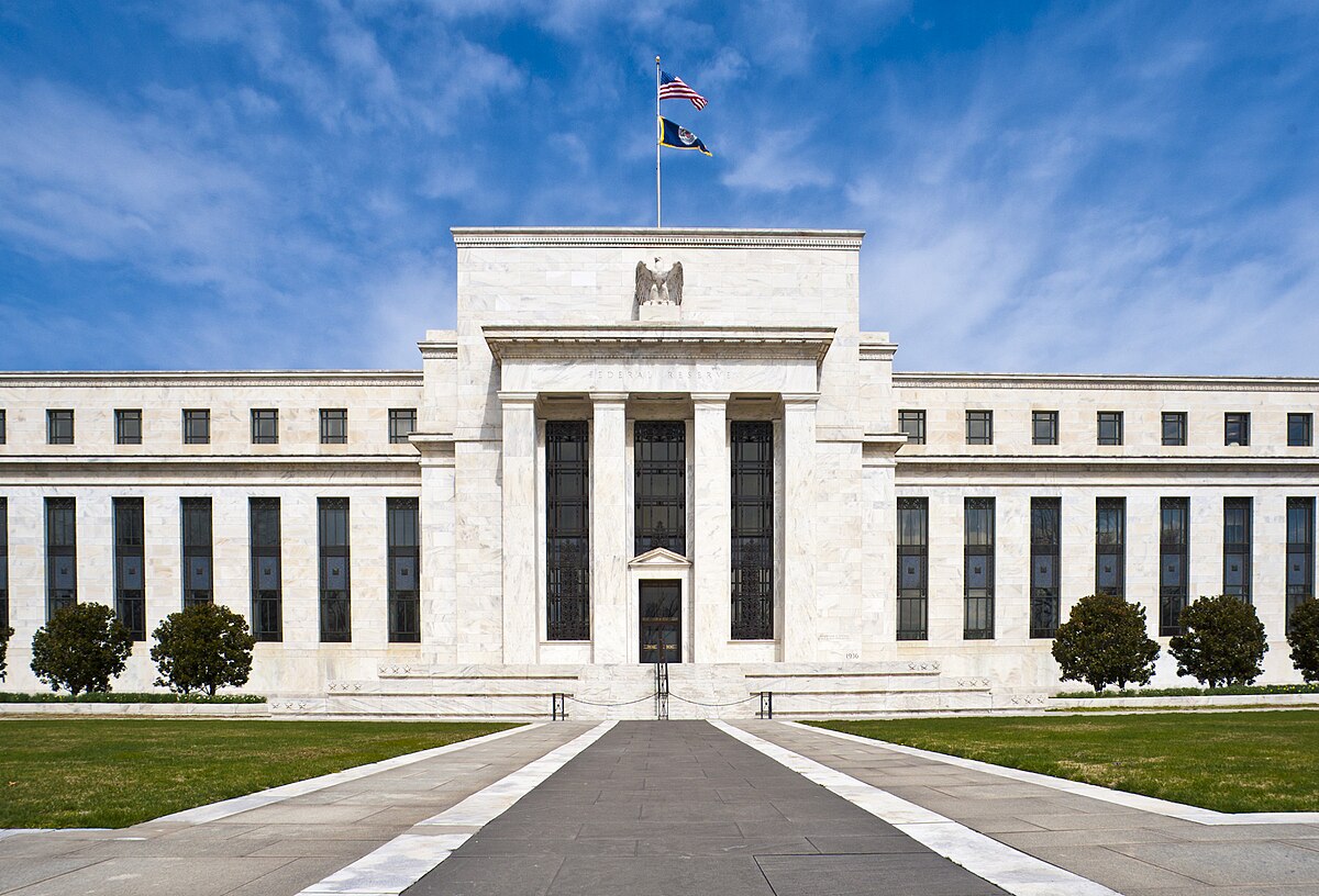Exterior view of the Marriner S. Eccles Federal Reserve Board Building in Washington, D.C., a large neoclassical structure with columns and a prominent entrance. The American flag and another flag are flying above the building.