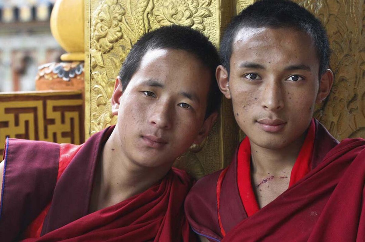 Two young men, dressed in red monastic robes, sit together in Bhutan, looking towards the viewer. They appear to be monks, with one resting his head on the other's shoulder.