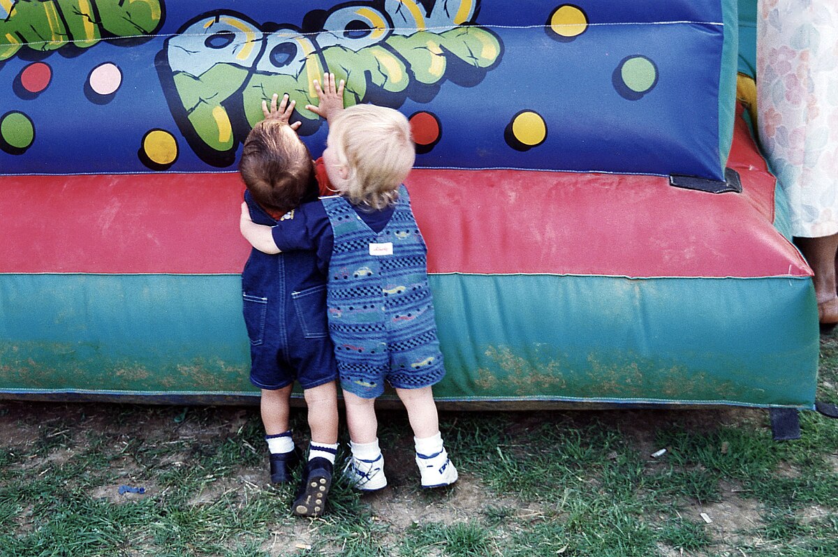 Two young children, seen from behind, stand close together and look at a colorful inflatable bounce house at a carnival. One child wears blue overalls and the other wears denim overalls.