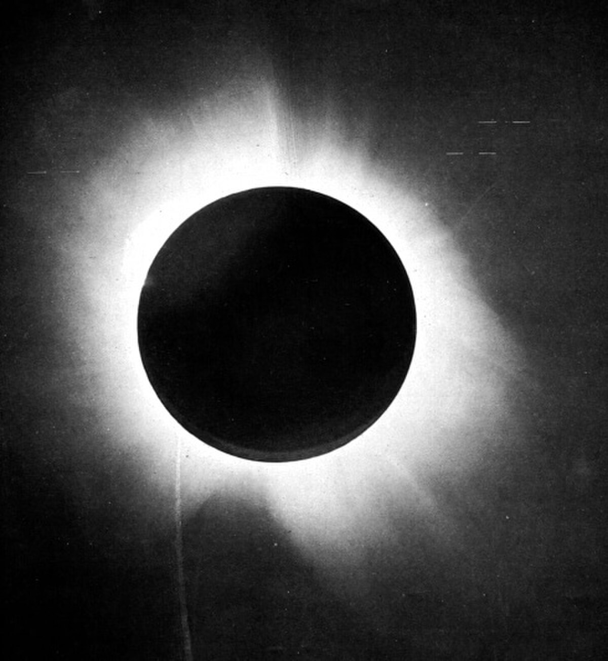 Black and white photograph of the 1919 total solar eclipse, showing the dark silhouette of the Moon obscuring the Sun, with the Sun's bright corona visible around its edges. Stars are faintly visible in the darkened sky.