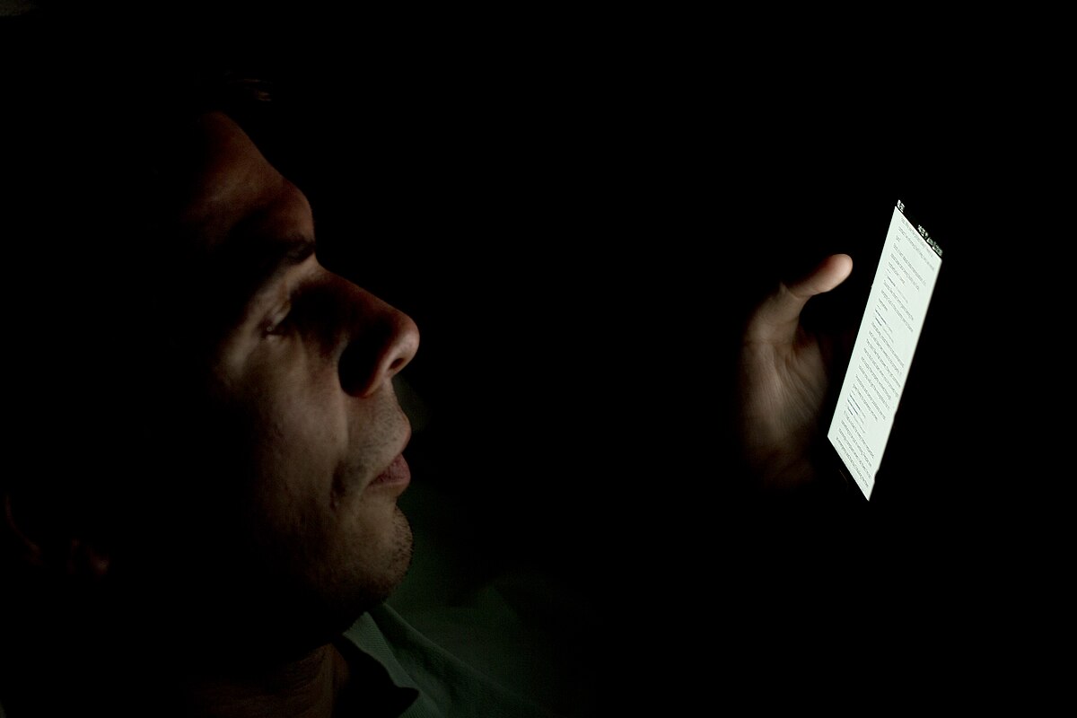 A person is shown in profile in a dark room, looking intently at the bright screen of a smartphone held in their hand. The light from the screen illuminates part of their face.