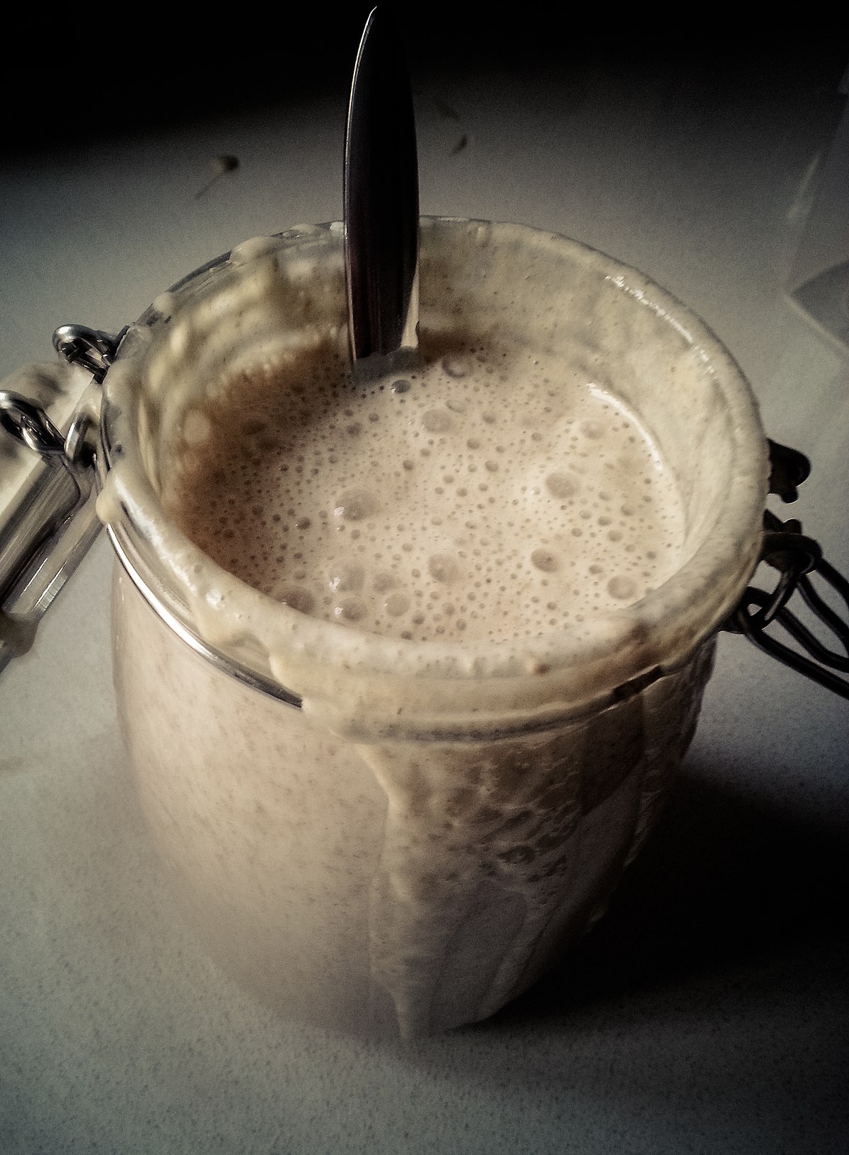 A glass jar filled with recently refreshed sourdough starter, showing a frothy, bubbly texture, with a spoon resting inside and some starter spilled over the rim.
