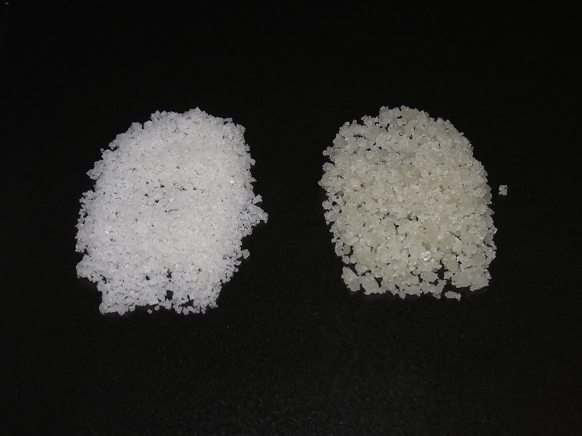Comparison of fleur de sel (left) and coarse salt (right) from the salterns of Guérande, displayed on a black surface. The fleur de sel appears finer and whiter, while the coarse salt has larger, more irregular crystals and a slightly off-white hue.