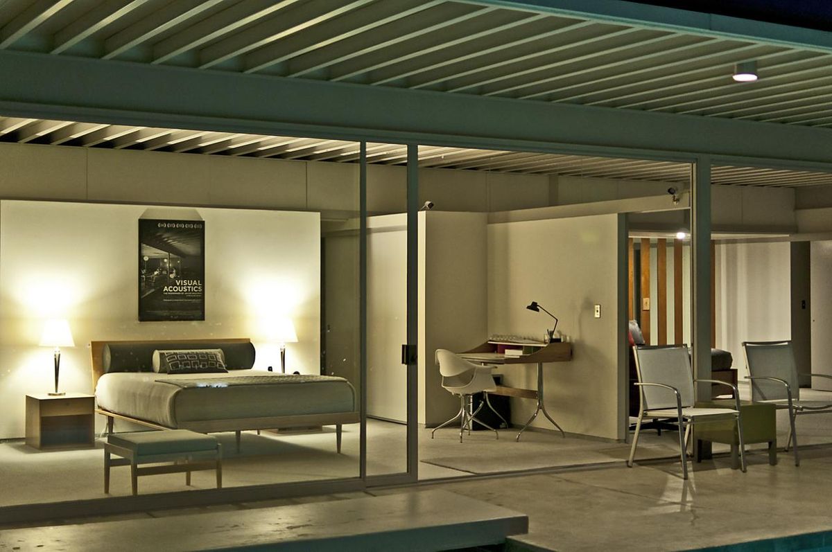 Interior view of the Stahl House (Case Study House 22) at dusk, showing a bedroom and an adjacent study area through its glass walls. The minimalist interior features a bed, desk, and chairs, with a poster on the wall and soft lighting illuminating the space.