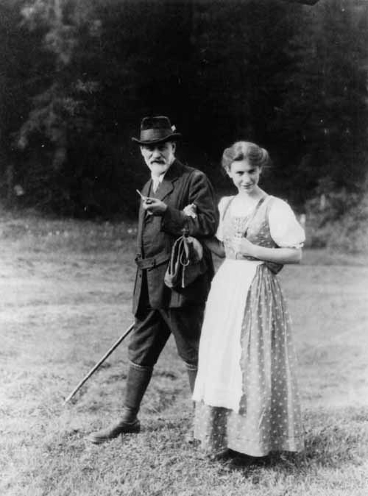 Anna Freud with her father Sigmund Freud in 1913, walking outdoors. Sigmund Freud is on the left, holding a walking stick and a bag, while Anna Freud is on the right, wearing a light-colored dress and apron.
