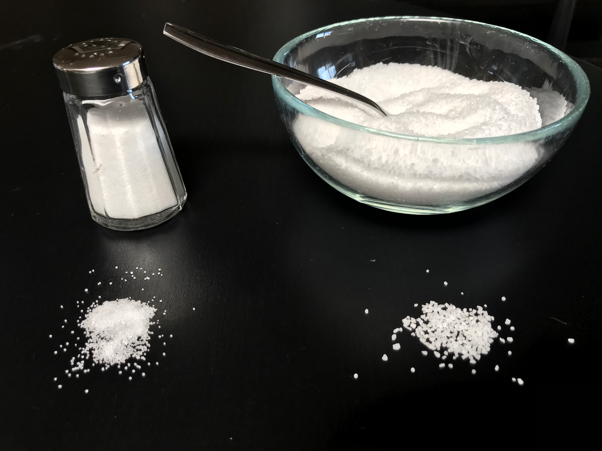 A comparison of table salt (left) and kosher salt (right) shows their different crystal sizes and shapes. Table salt is in a shaker and a small pile, while kosher salt is in a glass bowl with a spoon and a larger pile.