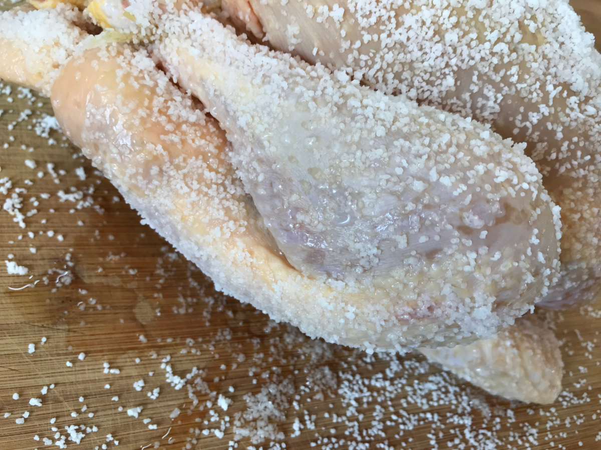 A whole chicken is covered in kosher salt for dry brining, with visible moisture extracted after one hour. The chicken rests on a wooden cutting board, and individual salt crystals are clearly visible.
