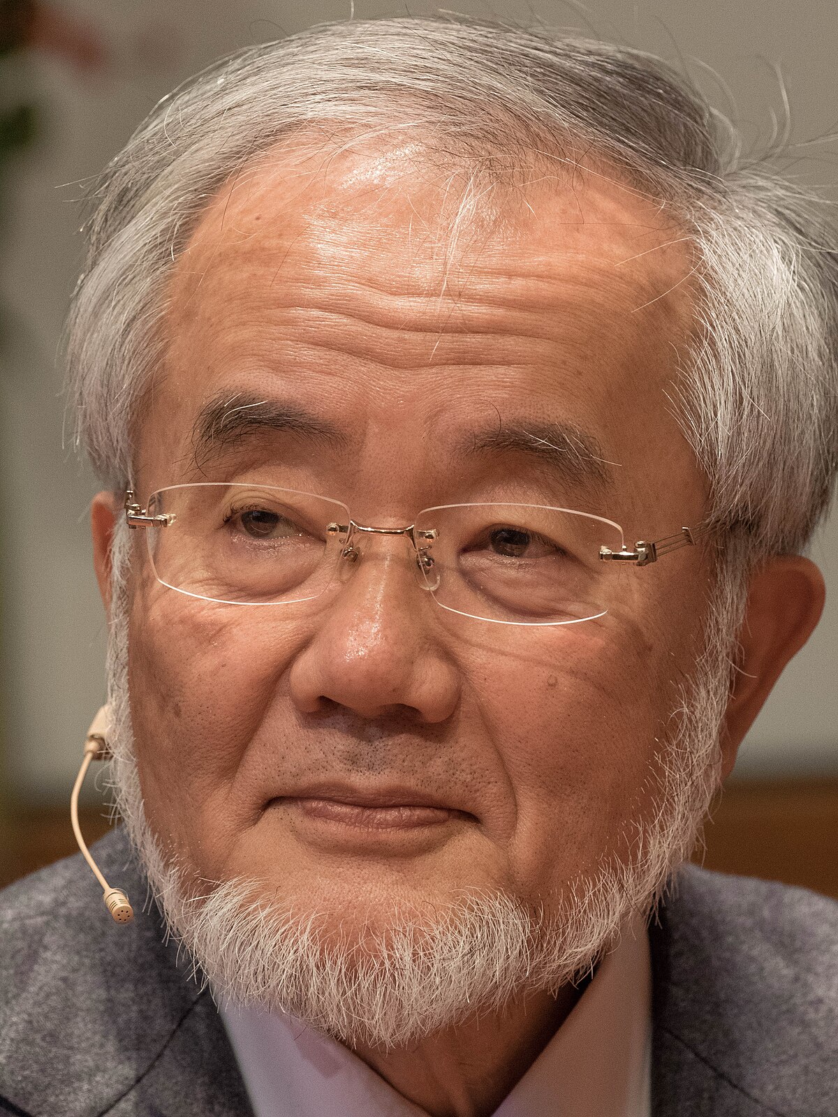 Portrait of Yoshinori Ohsumi, a Nobel Prize laureate, shown from the chest up, wearing glasses and a grey jacket, with a microphone near his right ear.
