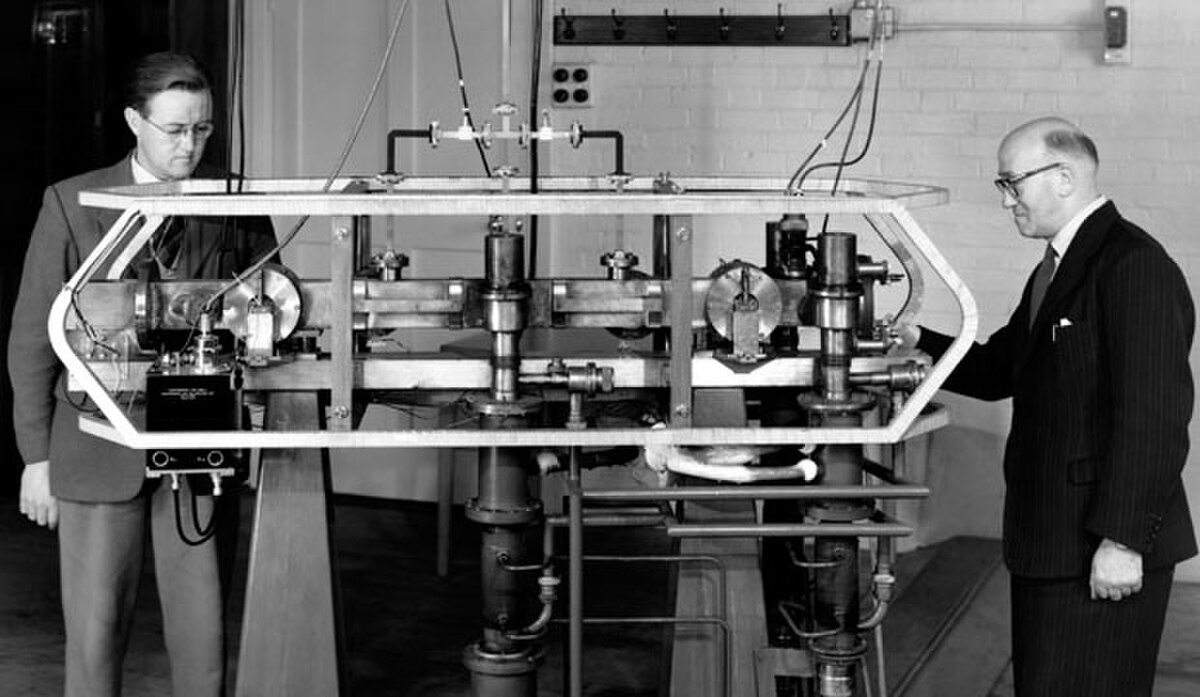 Louis Essen (right) and Jack Parry (left) standing next to the world's first caesium-133 atomic clock in 1955. The large, complex scientific instrument is composed of metal frames, tubes, and various components, reflecting early atomic clock technology.