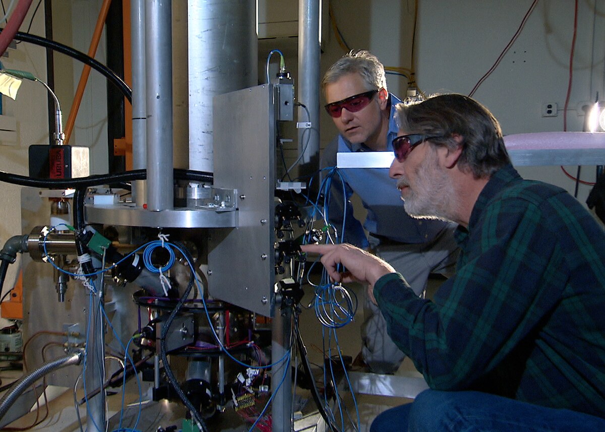 NIST physicists Steve Jefferts (foreground) and Tom Heavner (background) working with the NIST-F2 caesium fountain atomic clock. They are wearing safety glasses and examining the intricate components of the large, complex scientific apparatus.
