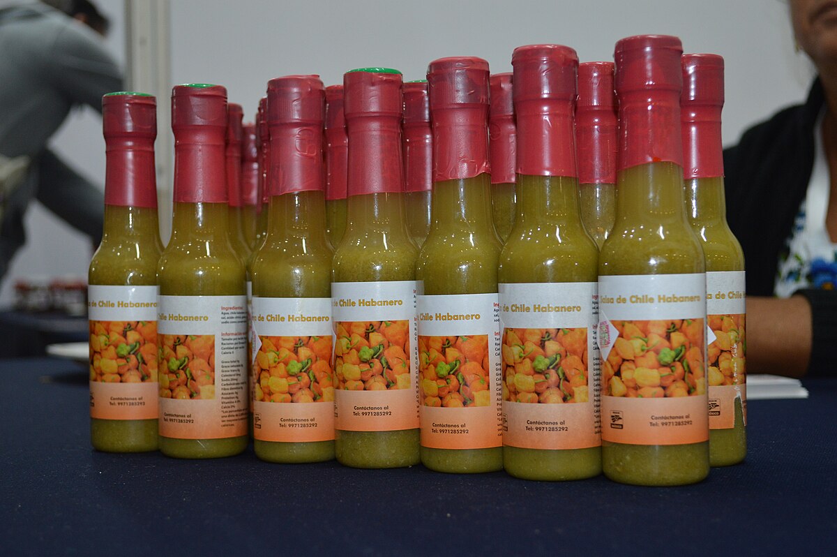 Multiple bottles of habanero hot sauce from Flor de Lirio, an Indigenous cooperative in Peto Municipality, Yucatán, are displayed on a table. Each bottle features a label with an image of diced habanero peppers and text in Spanish.