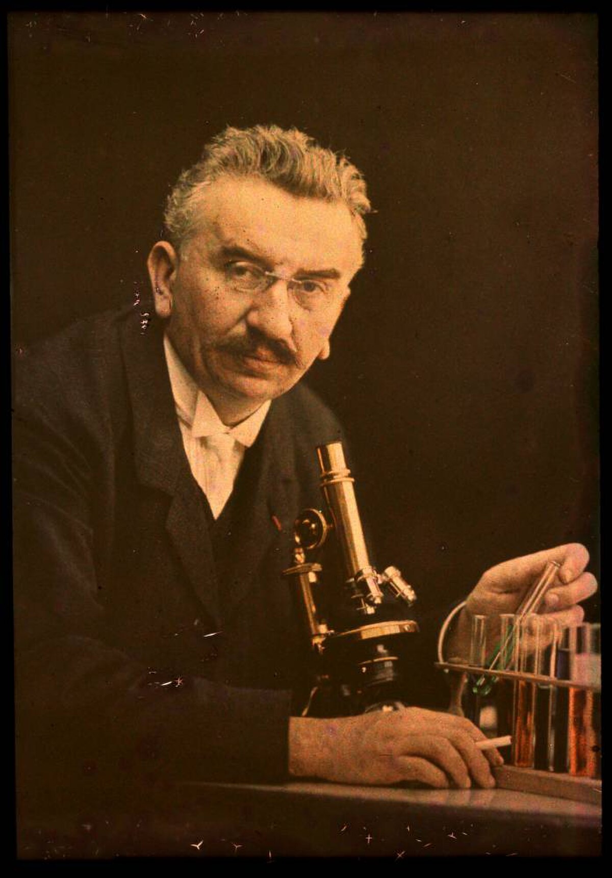 A colorized photograph of Louis Lumière, wearing glasses and a suit, seated at a table with a microscope and several test tubes in a rack. He is looking towards the viewer while holding a test tube.