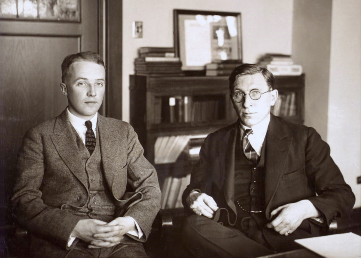 Black and white photograph of Frederick Banting (right) and Charles H. Best (left) seated in an office, taken around 1924. Both men are dressed in suits and ties, looking towards the camera.