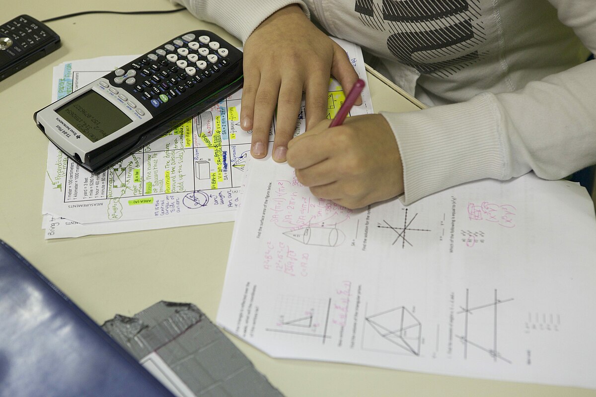 A student works on math problems with a graphing calculator and a pencil. The graphing calculator is a TI-84 Plus, and the student is writing on a worksheet with various geometric and algebraic problems.