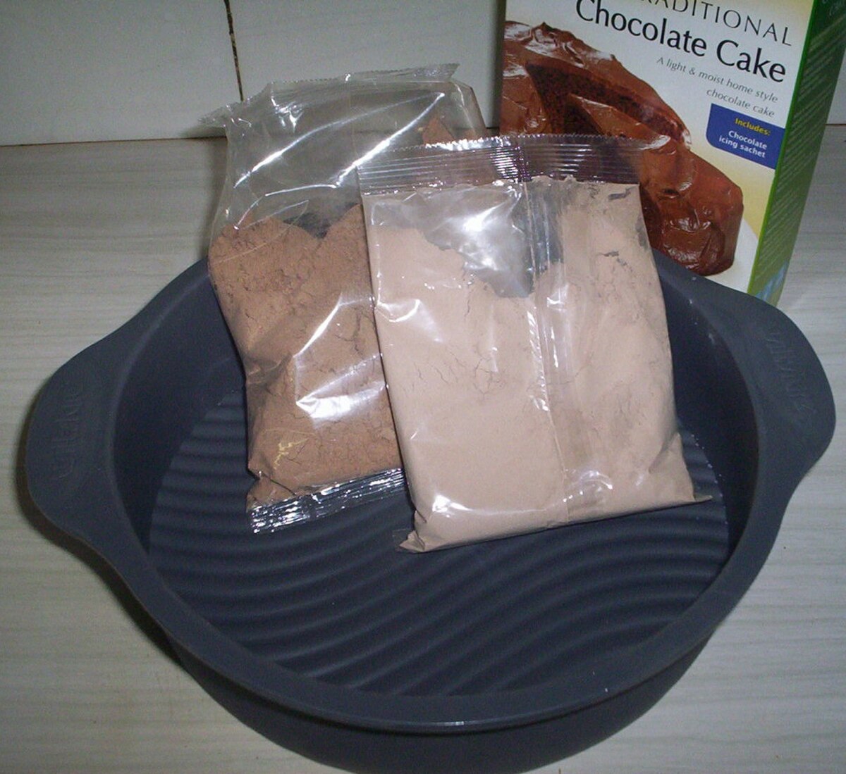 A cake mix in plastic packets, placed inside a gray baking pan, with a box for "Traditional Chocolate Cake" visible in the background. The packets contain dry ingredients for baking.
