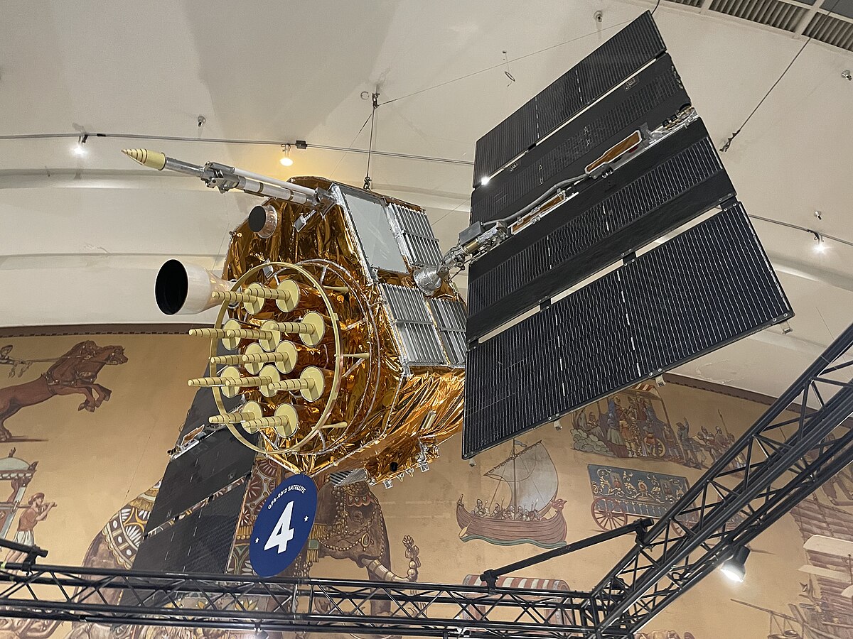 A qualification vehicle for GPS Block II is on display at the San Diego Air & Space Museum, showcasing its intricate design with numerous antennas and large solar panels. A sign with the number "4" is visible in the foreground.