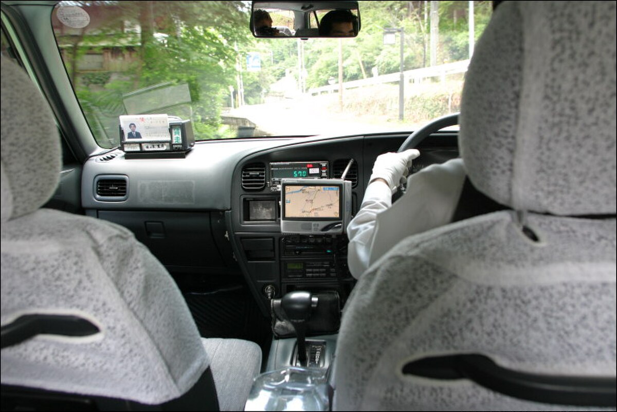 The interior of a taxi in Kyoto, Japan, shows a GPS navigation system mounted on the dashboard, with the driver's hands on the steering wheel. The rear view mirror reflects the road behind, and the seats are covered with white protective covers.