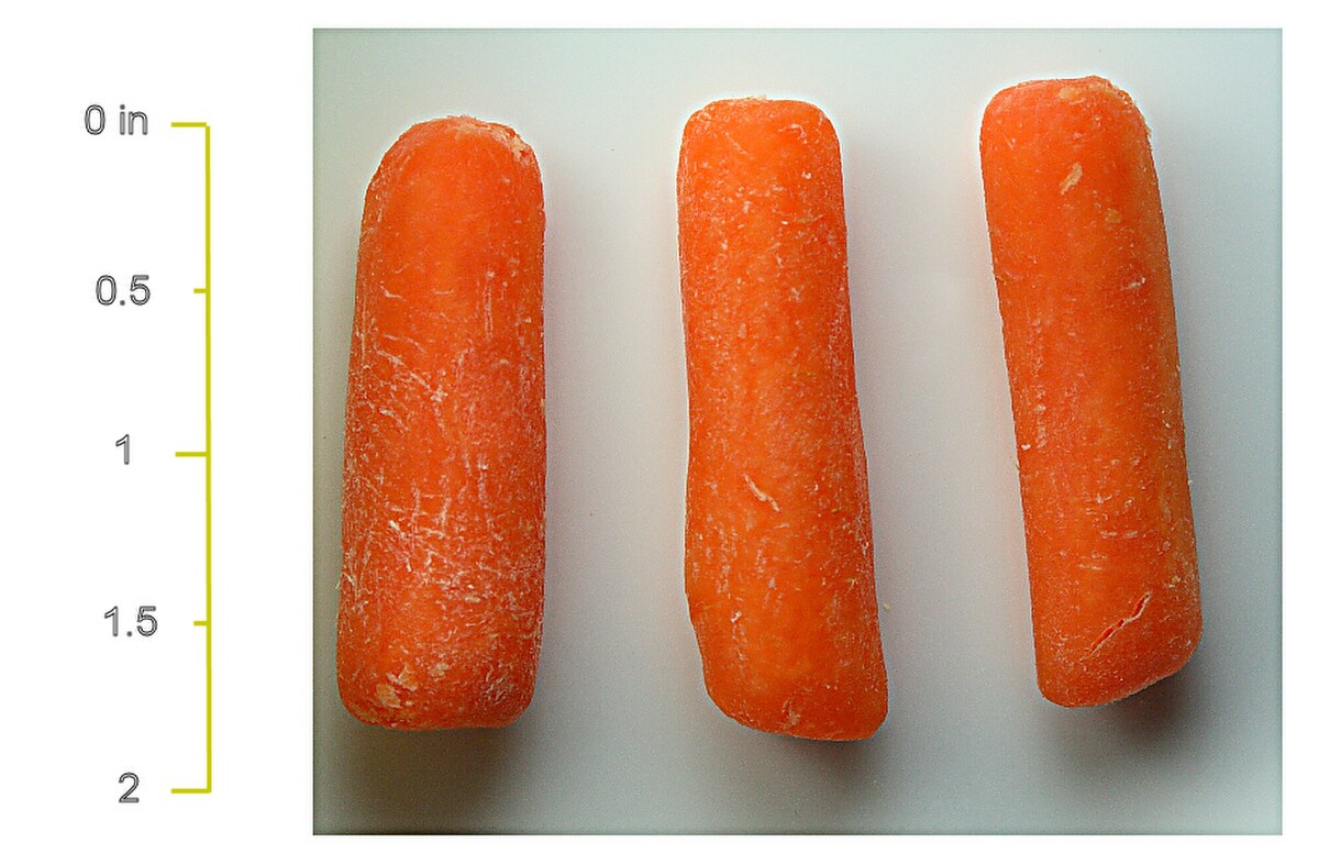 Three baby-cut carrots are arranged horizontally on a white surface, with a vertical ruler on the left indicating measurements in inches from 0 to 2. Each carrot is approximately 1.5 inches long.