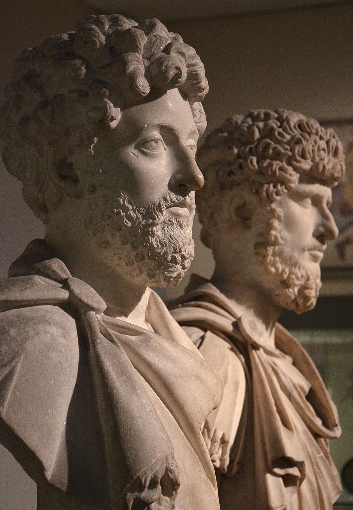 Two marble busts of Roman co-emperors, Marcus Aurelius (left) and Lucius Verus (right), displayed at the British Museum. Both busts depict men with curly hair and beards, draped in togas.