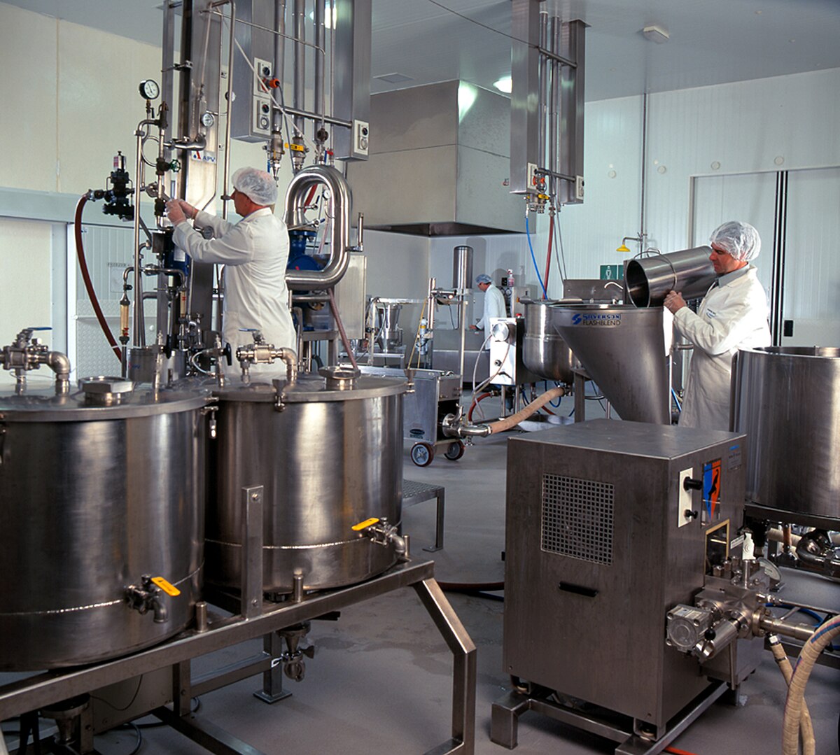 Two food scientists in white lab coats and hairnets work in a laboratory in Australia, surrounded by large stainless steel processing equipment. One scientist is adjusting controls on a tall apparatus, while the other is pouring material into a hopper.