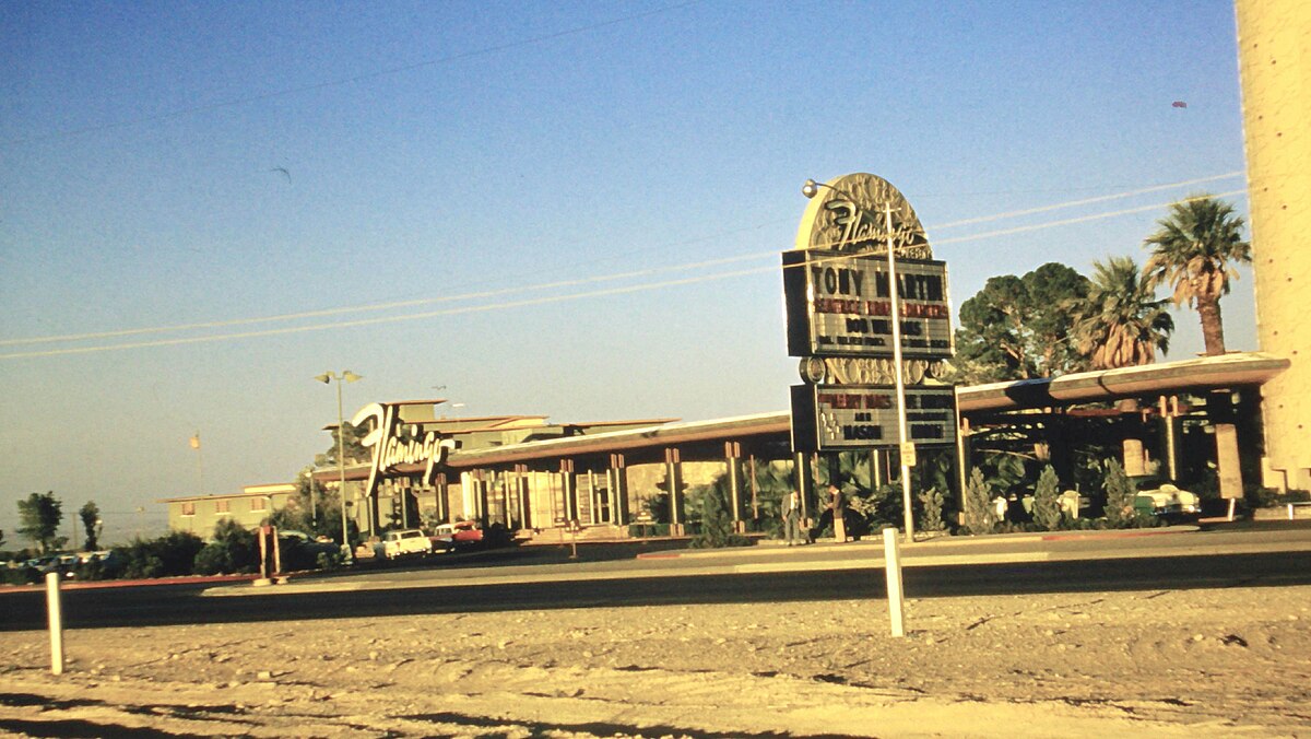 The Flamingo resort in Las Vegas in 1957, showing the original structure with a large vertical Flamingo sign. The building is long and low-slung, with palm trees in the foreground.