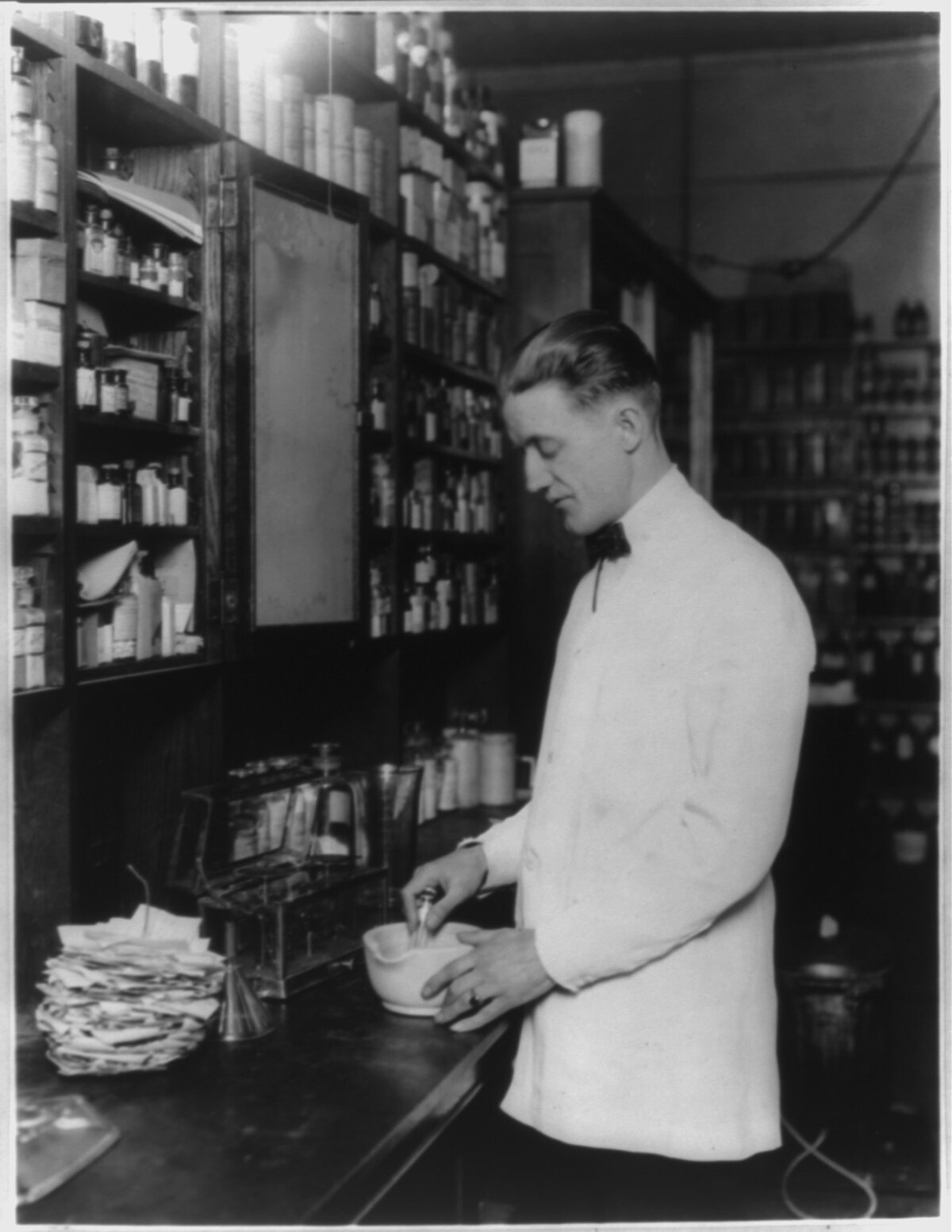 A black and white photograph from approximately 1923, showing a male pharmacist compounding a medication using a mortar and pestle in a drug store. Shelves filled with various bottles and containers are visible in the background.