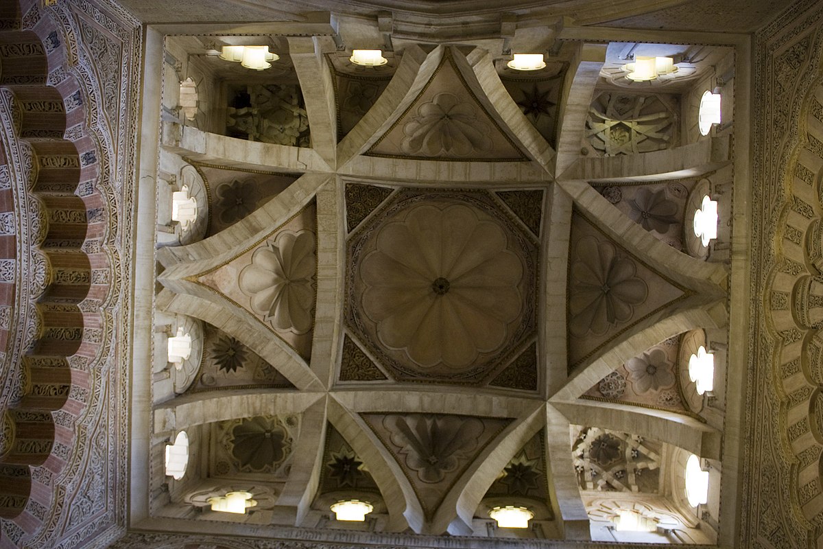An upward view of a complex rib vault ceiling in the Capilla Real (Royal Chapel) of the Mezquita-Cathedral of Córdoba, Spain. The ceiling features a square base with intersecting ribs forming a star-like pattern and smaller decorative elements within the vaulted sections.