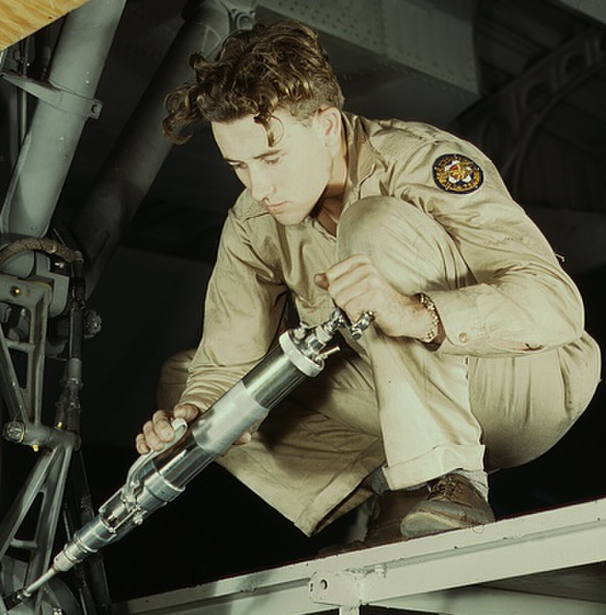 A Consolidated Aircraft hydraulic mechanic greasing the landing gear of a transport aircraft. The mechanic, wearing a light-colored uniform with a patch on his right arm, is crouched and focused on the task, holding a grease gun.