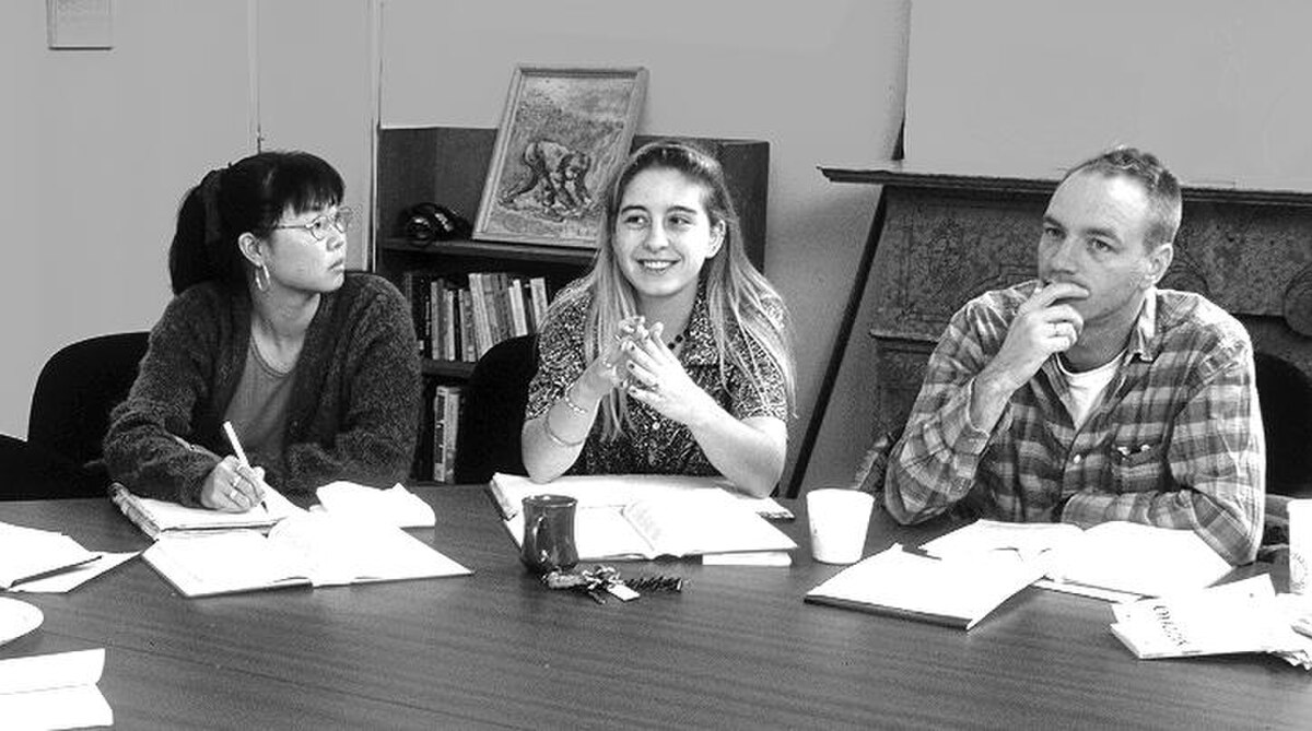 Black and white photograph of three Shimer College students engaged in a discussion around a table, likely as part of their core curriculum. The students are looking at each other and appear to be taking notes or reading texts.