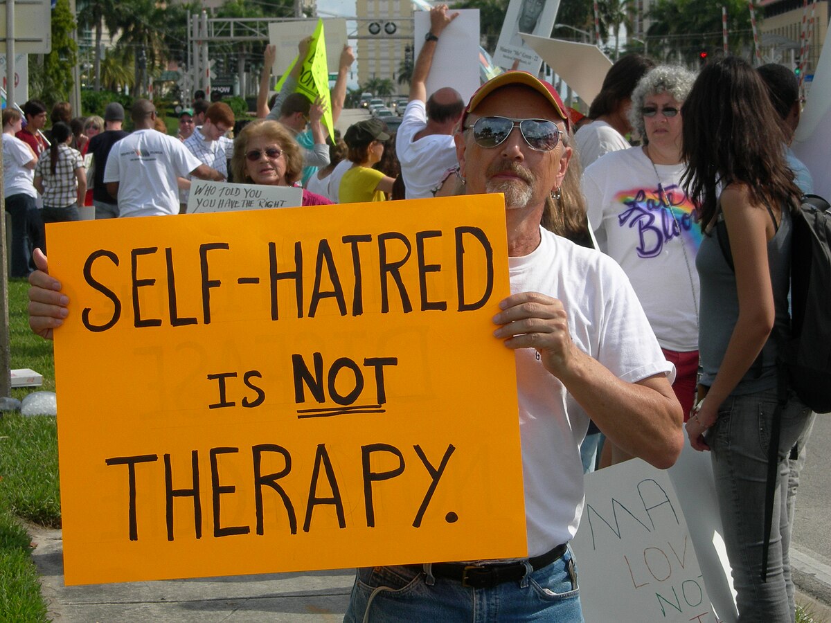 A man holding a large orange sign that reads "SELF-HATRED IS NOT THERAPY" during a protest against conversion therapy in 2009. The protest highlights the criticisms surrounding conversion therapy and its association with sexual repression, shame, and guilt among gay, lesbian, and bisexual individuals.