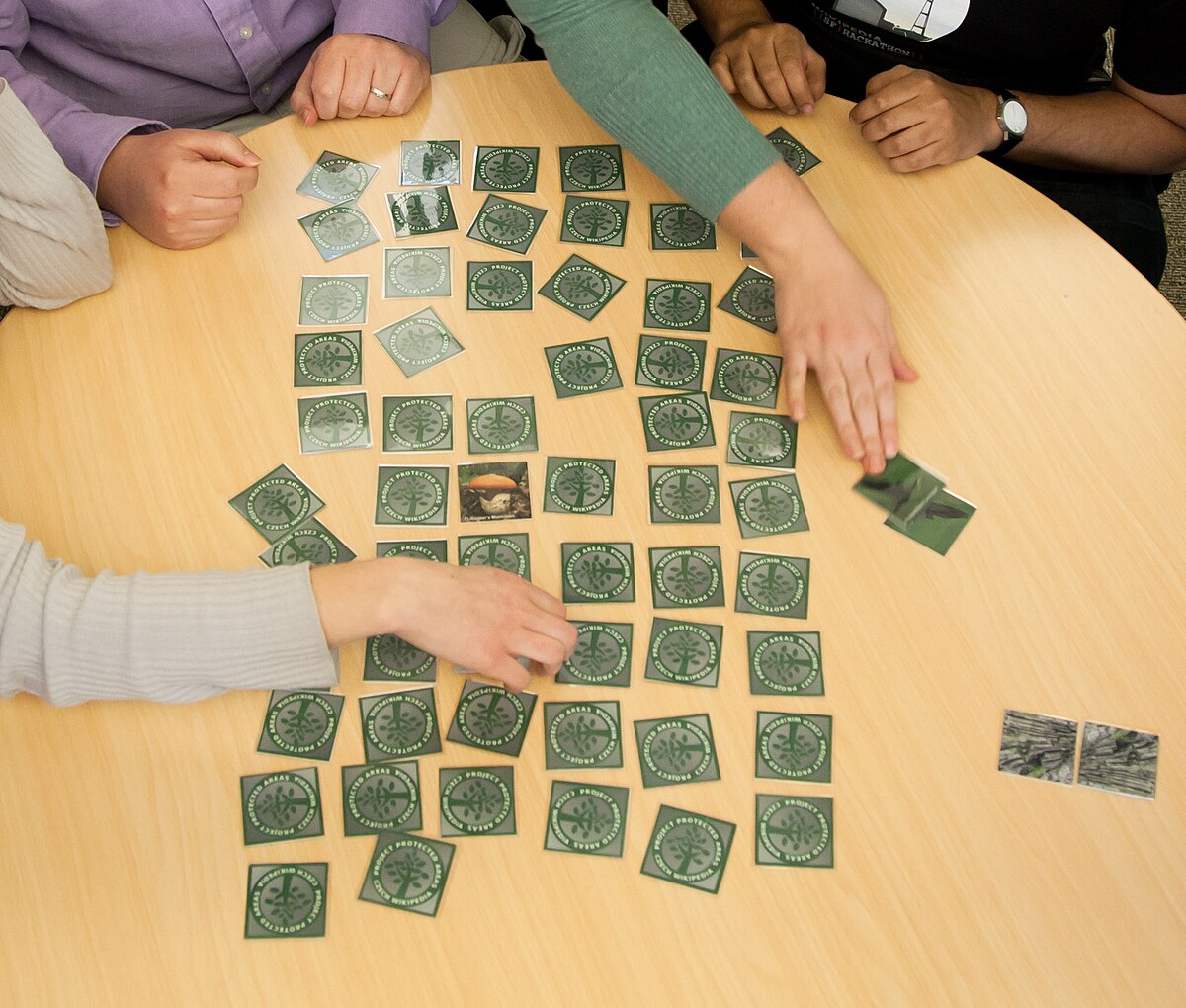 Several people play a card game of Concentration, with numerous green square cards spread across a round wooden table. Hands reach for cards, and some matching pairs have been removed from the main playing area.