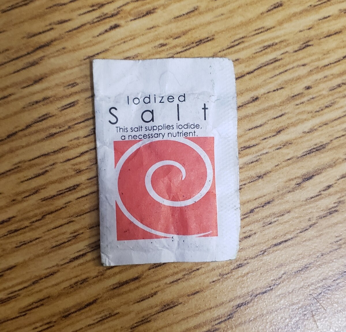 A small white packet of iodized salt with a red spiral logo. The text on the packet reads "Iodized Salt This salt supplies iodide, a necessary nutrient."