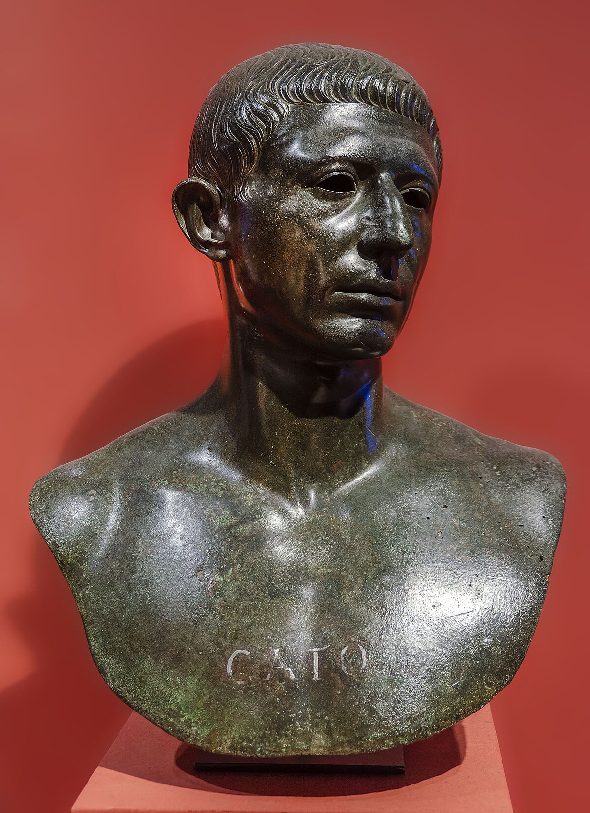 A bronze bust of Cato the Younger is displayed against a red background. The name "CATO" is inscribed on the base of the bust.