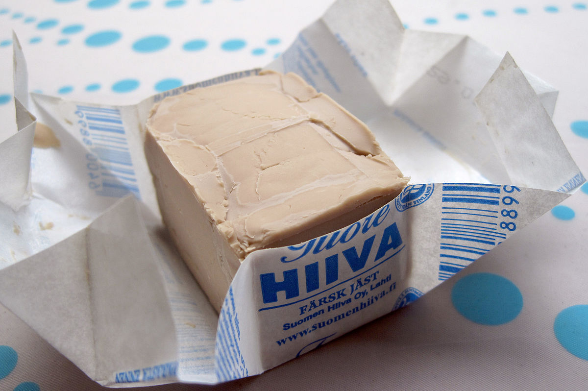 A block of compressed fresh yeast, partially unwrapped from its paper packaging. The packaging shows text in Finnish and Swedish, including "Tuore HIIVA" (Fresh Yeast) and "FÄRSK JÄST."