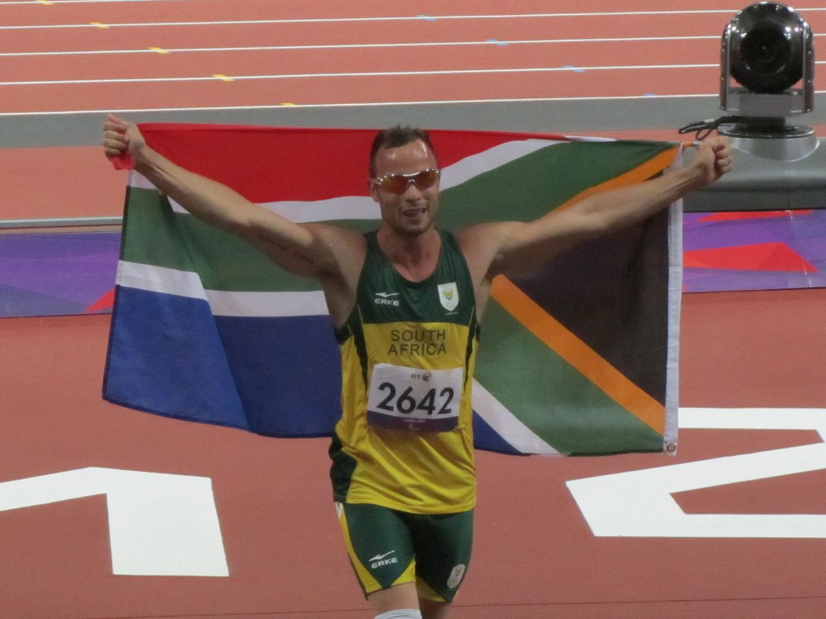 Oscar Pistorius, wearing a green and yellow South Africa uniform with the number 2642, holds a large South African flag above his head on a running track. He is smiling and wearing orange sunglasses.