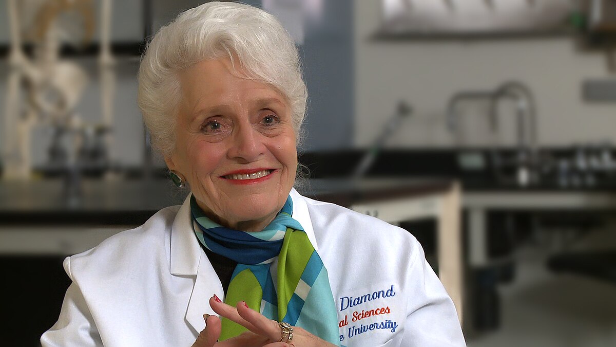 Portrait of Dr. Marian Diamond smiling while wearing a white lab coat and a colorful scarf. The lab coat has "Diamond Medical Sciences University" embroidered on it.