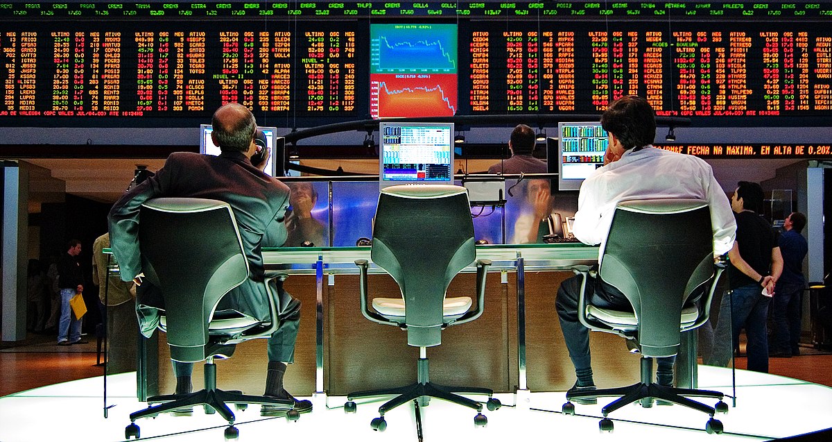 Two traders are seated at desks in front of multiple computer monitors displaying stock market data and charts at the Sao Paulo Stock Exchange. A large digital ticker board with numerous stock prices is visible in the background.