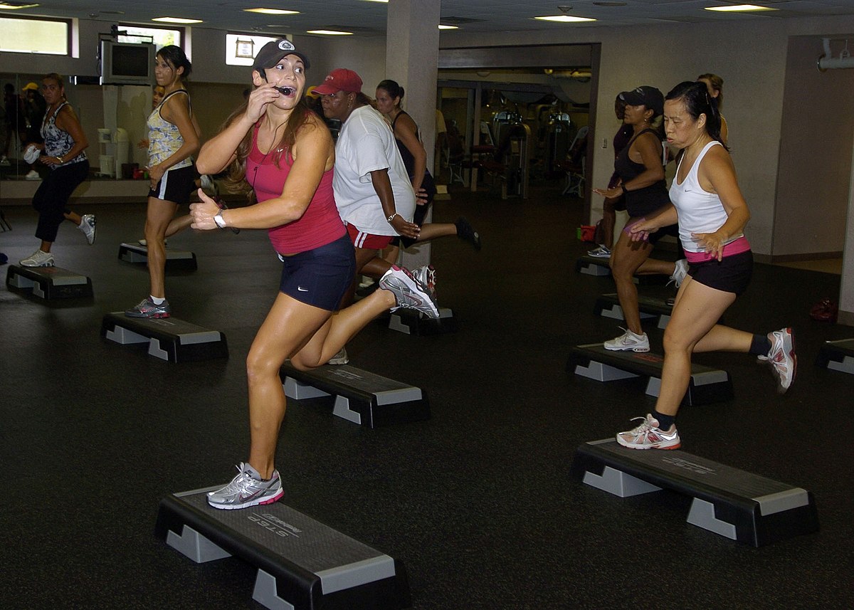 A step aerobics instructor leads a class of people performing exercises on step platforms in a gym. The instructor, in the foreground, wears a headset microphone and gives a thumbs-up gesture.