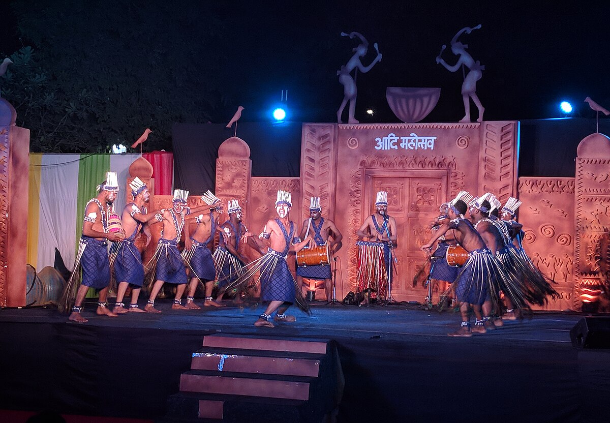 A group of Siddi tribal dancers are performing on a stage in Delhi, illuminated by stage lights. They are wearing traditional costumes and playing drums, with a decorated backdrop featuring figures and text in Hindi.