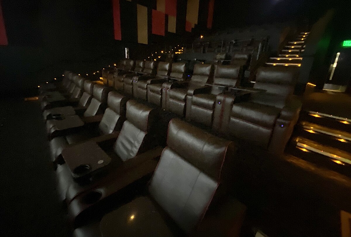 An interior view of a digital cinema theater featuring stadium seating with comfortable, reclining brown leather chairs, each equipped with a small table and cup holder.