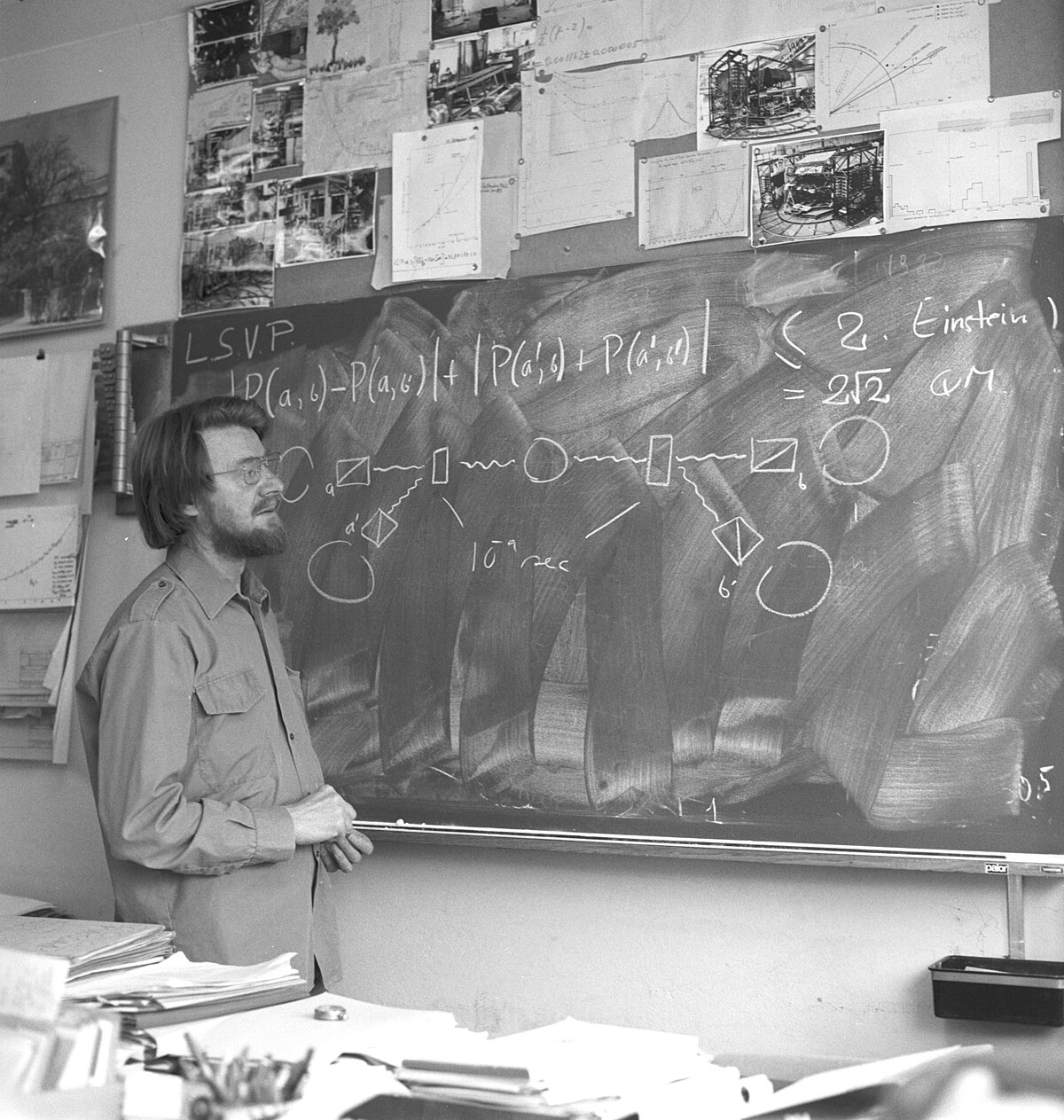 A black and white photograph shows John Stewart Bell discussing Bell's inequality at CERN in 1982, standing in front of a blackboard filled with physics equations and diagrams. The blackboard includes the expression "|D(a,c)-P(a,c)| + |P(a',c') + P(a',c')|" and "(2. Einstein) = 2√2 @M".