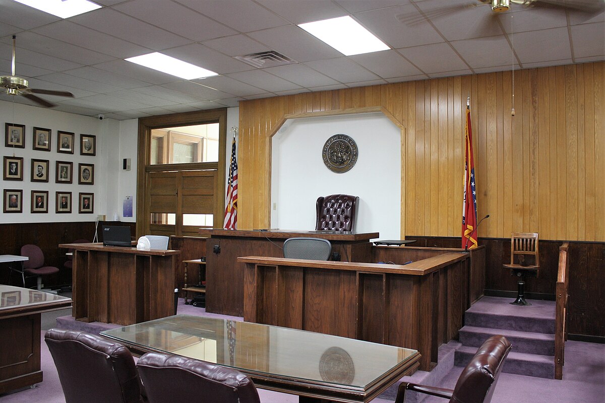 A courtroom inside the Boone County Courthouse in Boone County, Arkansas, featuring dark wood paneling, a judge's bench, and jury box. An American flag and an Arkansas state flag stand to the right of the judge's bench, and framed portraits line the left wall.